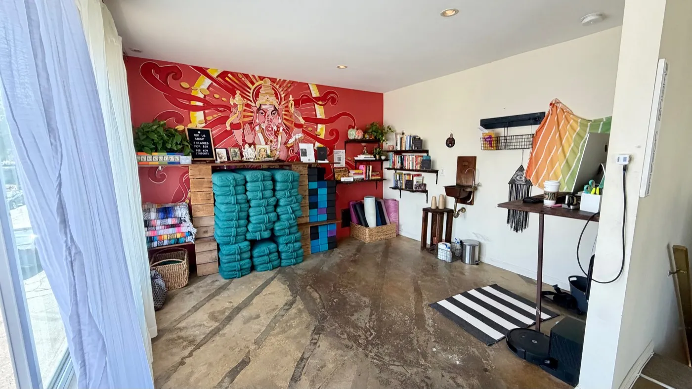 The prop room in full: the Ganesha mural and bolster stacks along the red wall, folded blankets and rolled mats to one side, bookshelves, a striped runner and a hanging towel rail opposite