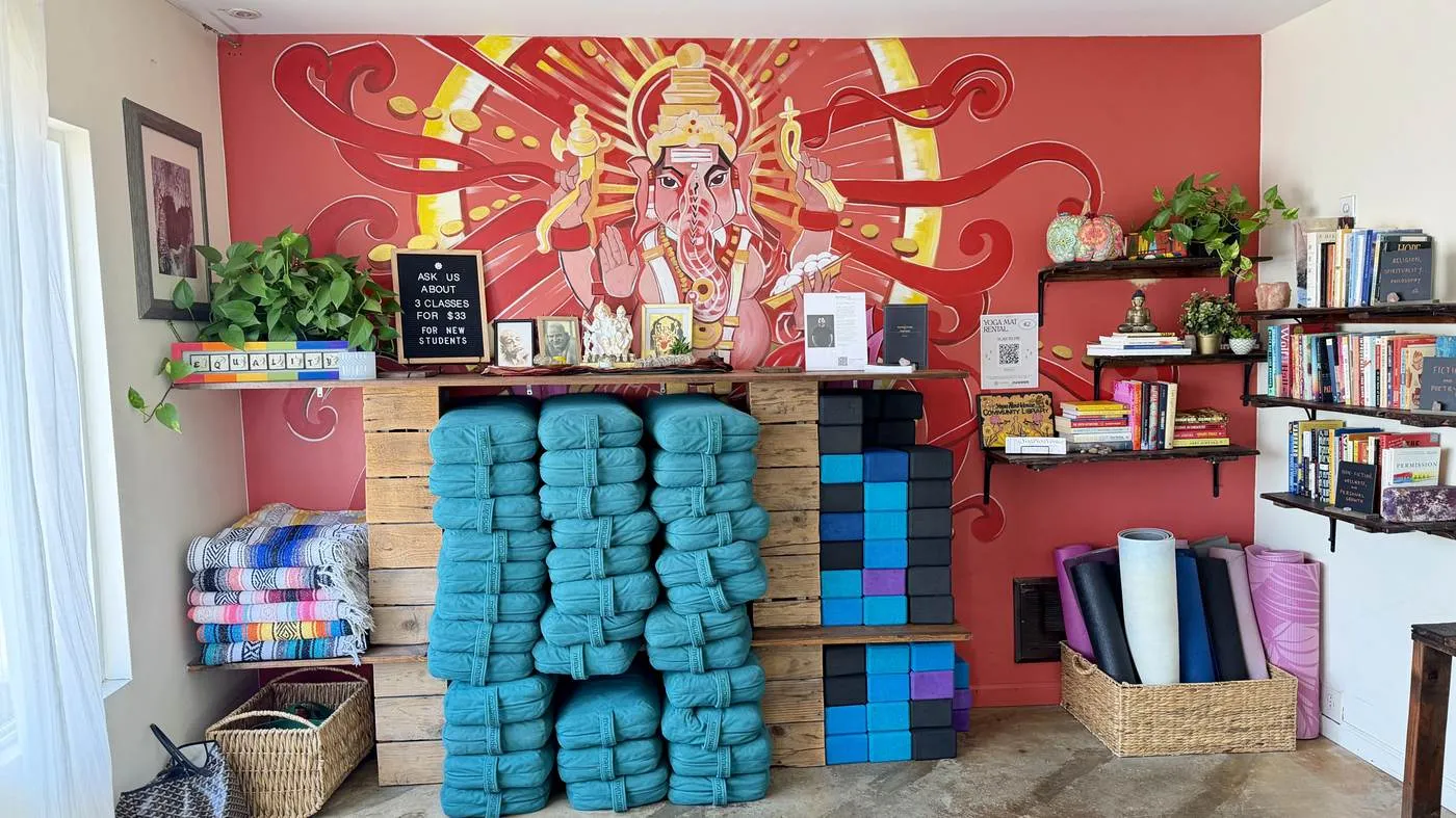 The prop wall: stacks of teal blankets and foam blocks on pallet shelving against a red wall painted with a Ganesha mural, rolled mats and bookshelves either side