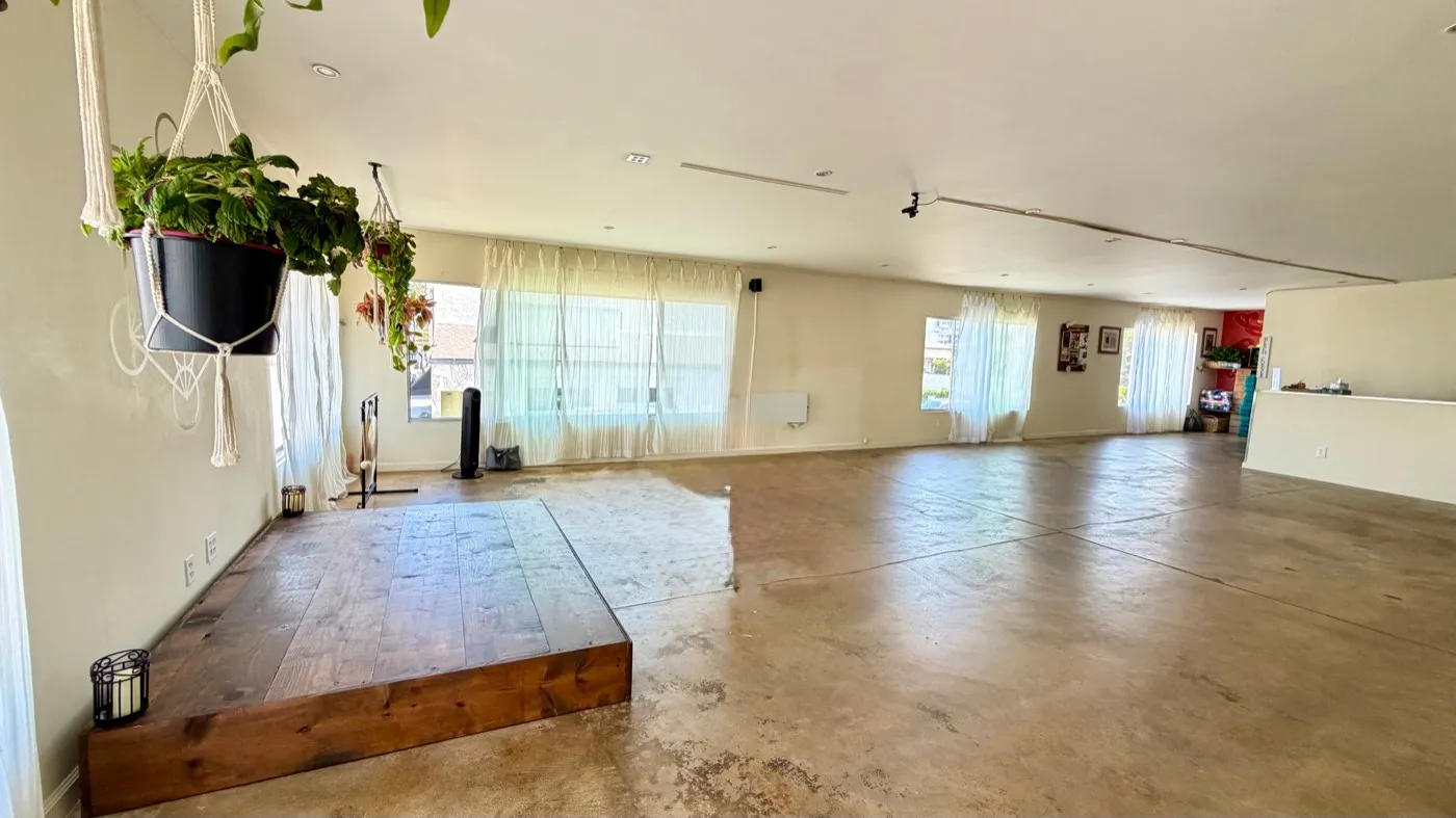 The practice room along its full length: the raised wooden platform in the foreground, sheer-curtained windows down the left side and the prop room visible at the far end