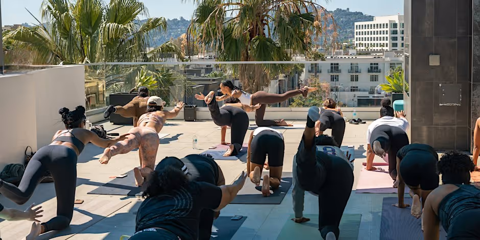 A rooftop class mid-flow on mats, several students balancing in warrior three, with palms and West Hollywood rooftops behind them